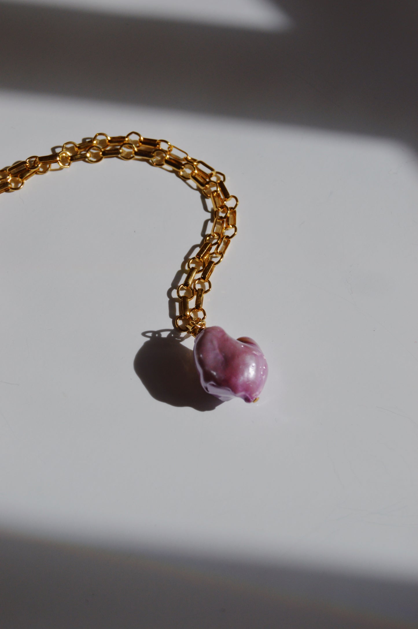 The Lavender Pearl Drop Necklace features a large, irregular pink freshwater pearl pendant on a 14k gold-filled chain, displayed on a white surface with subtle shadows.