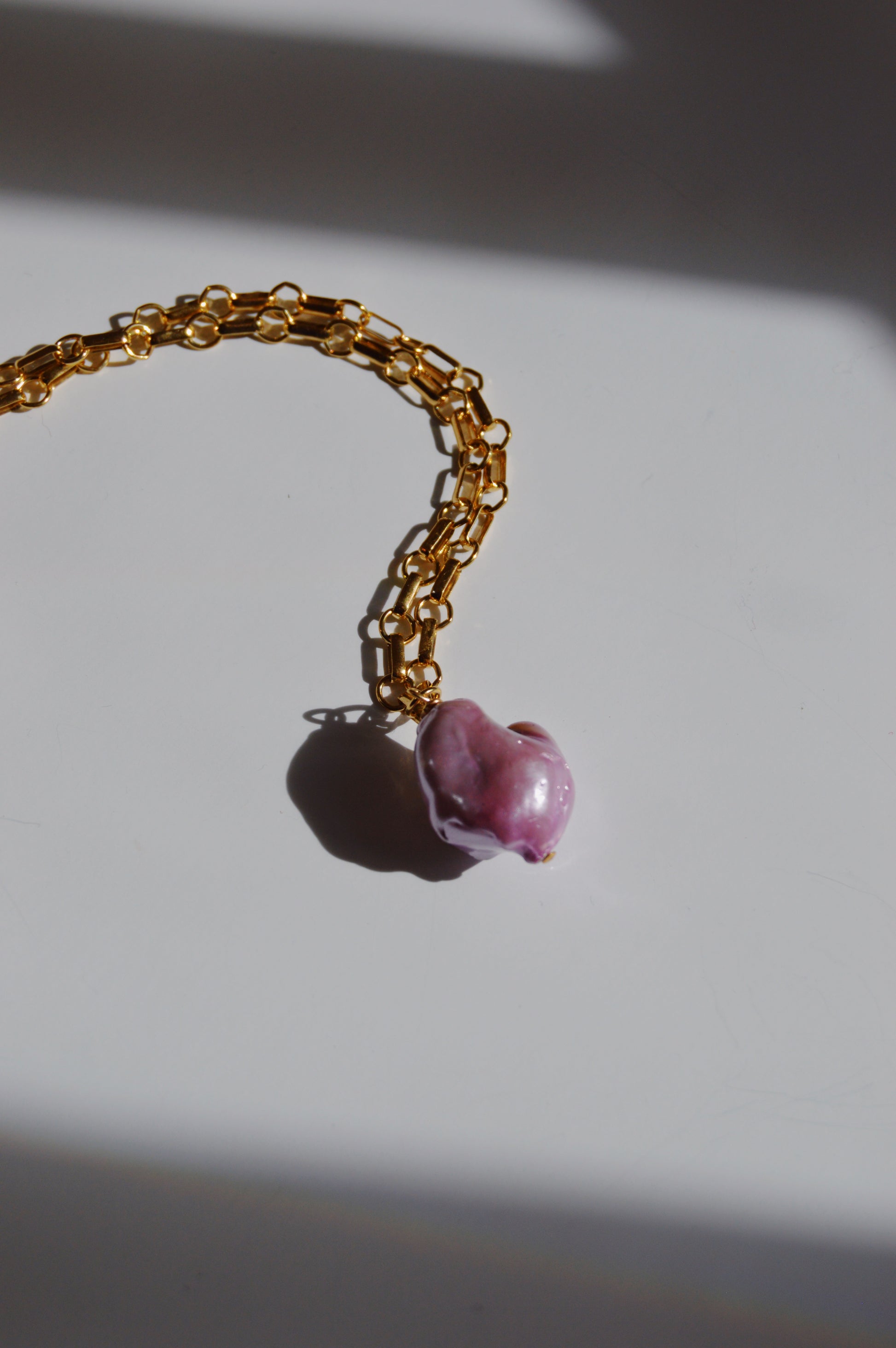 The Lavender Pearl Drop Necklace features a large, irregular pink freshwater pearl pendant on a 14k gold-filled chain, displayed on a white surface with subtle shadows.