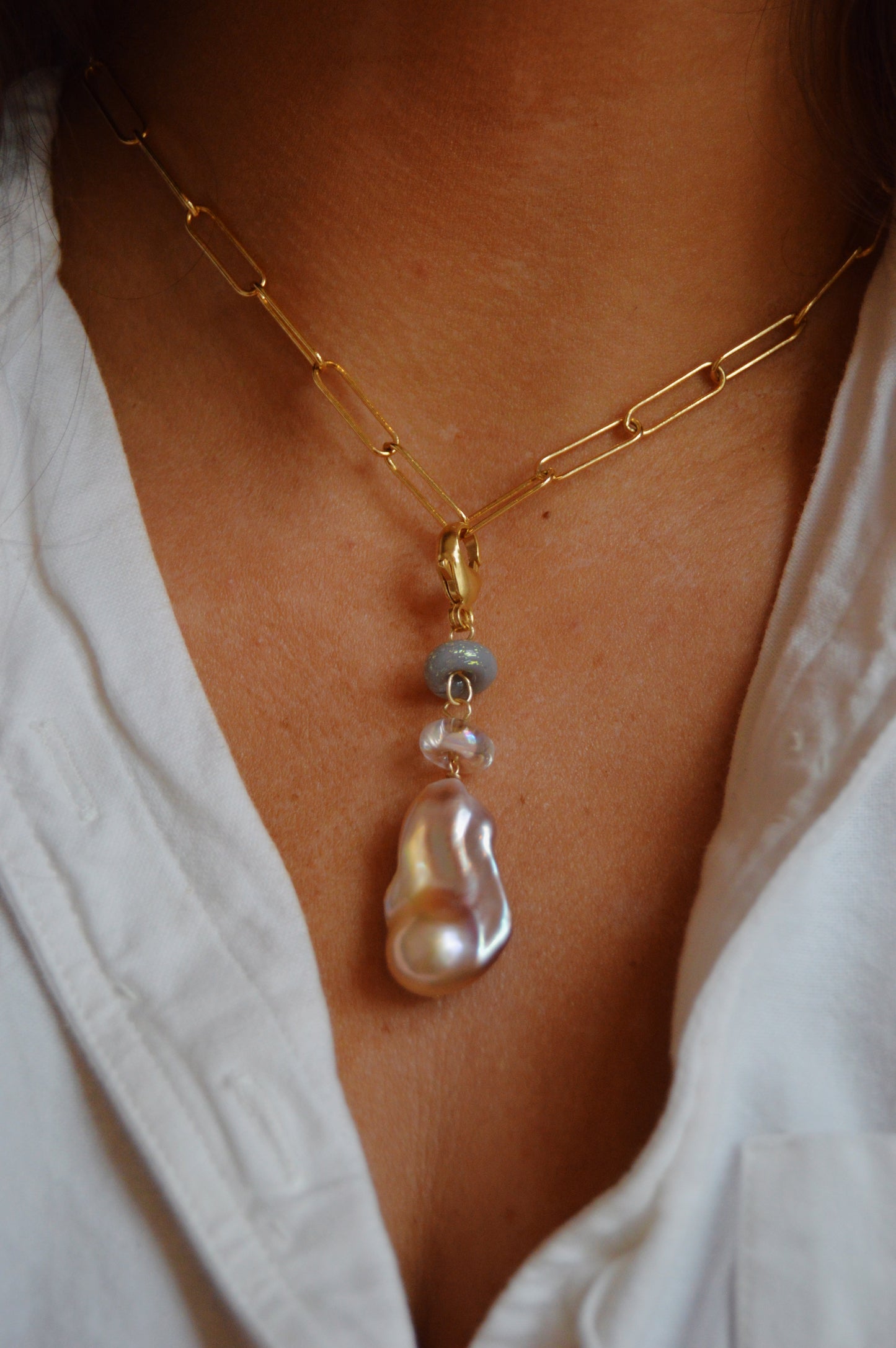 A close-up of the Glass and Pearl Charm necklace, featuring a gold paperclip chain with a large cultured freshwater pearl, blue handmade lampwork glass bead, and clear bead pendant, styled with a white shirt.