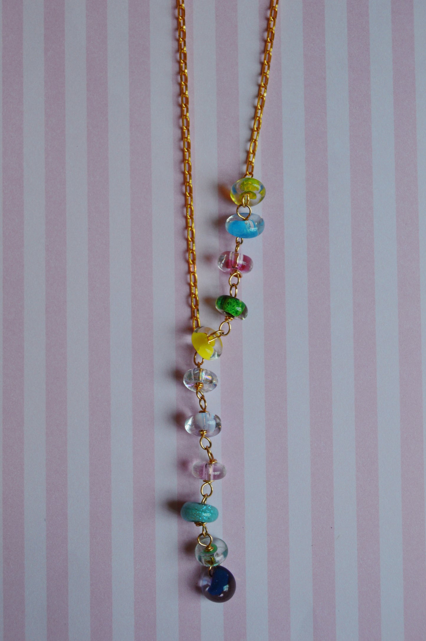 The Valentine Donut Lariat features a gold chain with colorful glass beads in blue, green, yellow, pink, and purple, displayed on a pastel pink and white striped background.
