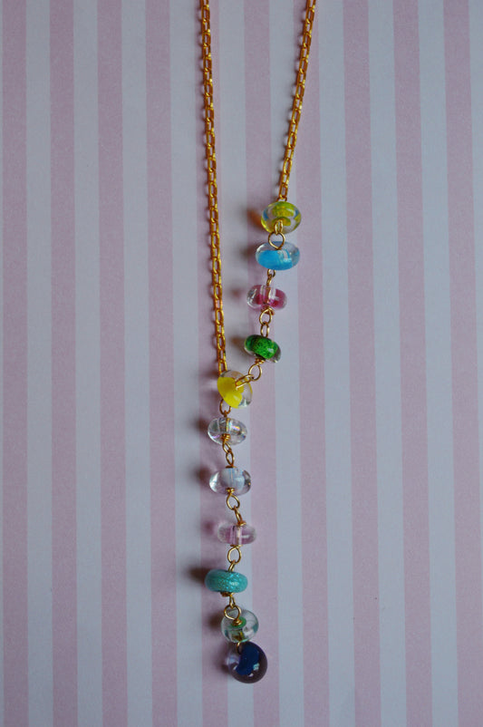 The Valentine Donut Lariat features a gold chain with colorful glass beads in blue, green, yellow, pink, and purple, displayed on a pastel pink and white striped background.