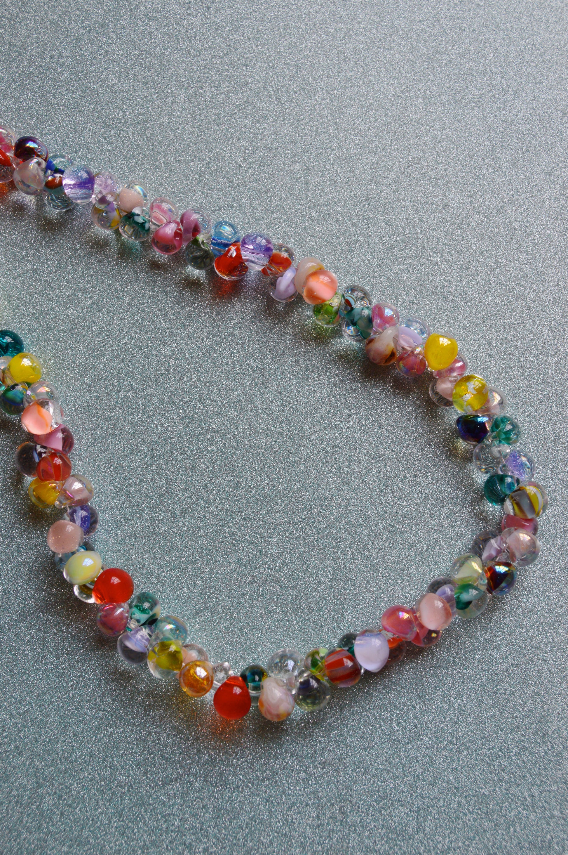 The Rainbow Bubble Glass Necklace features colorful glass beads in red, yellow, green, blue, and purple with gold filled findings, arranged on a textured light gray surface.