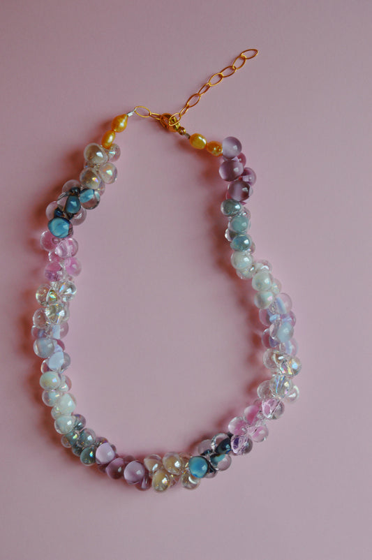 The Valentine Bubble Glass Necklace- Sweetheart features clusters of pastel pink, purple, blue, and white translucent beads on a gold chain with clasp, beautifully displayed against a pink background.