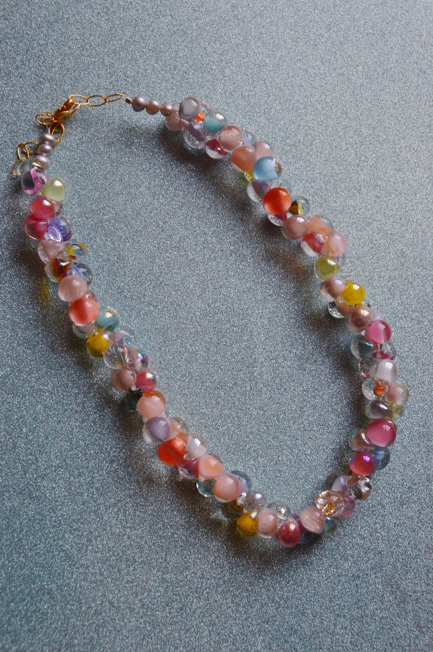 The Rainbow Bubble Glass Necklace features multi-colored, irregular glass beads and gold filled findings in pink, blue, yellow, and clear tones, shown on a textured gray surface.