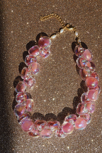 The Grand Bubble Glass Necklace-Shimmer Pink, featuring iridescent pink and clear glass beads, freshwater pearls, and a gold clasp, is displayed on a sparkling brown surface.