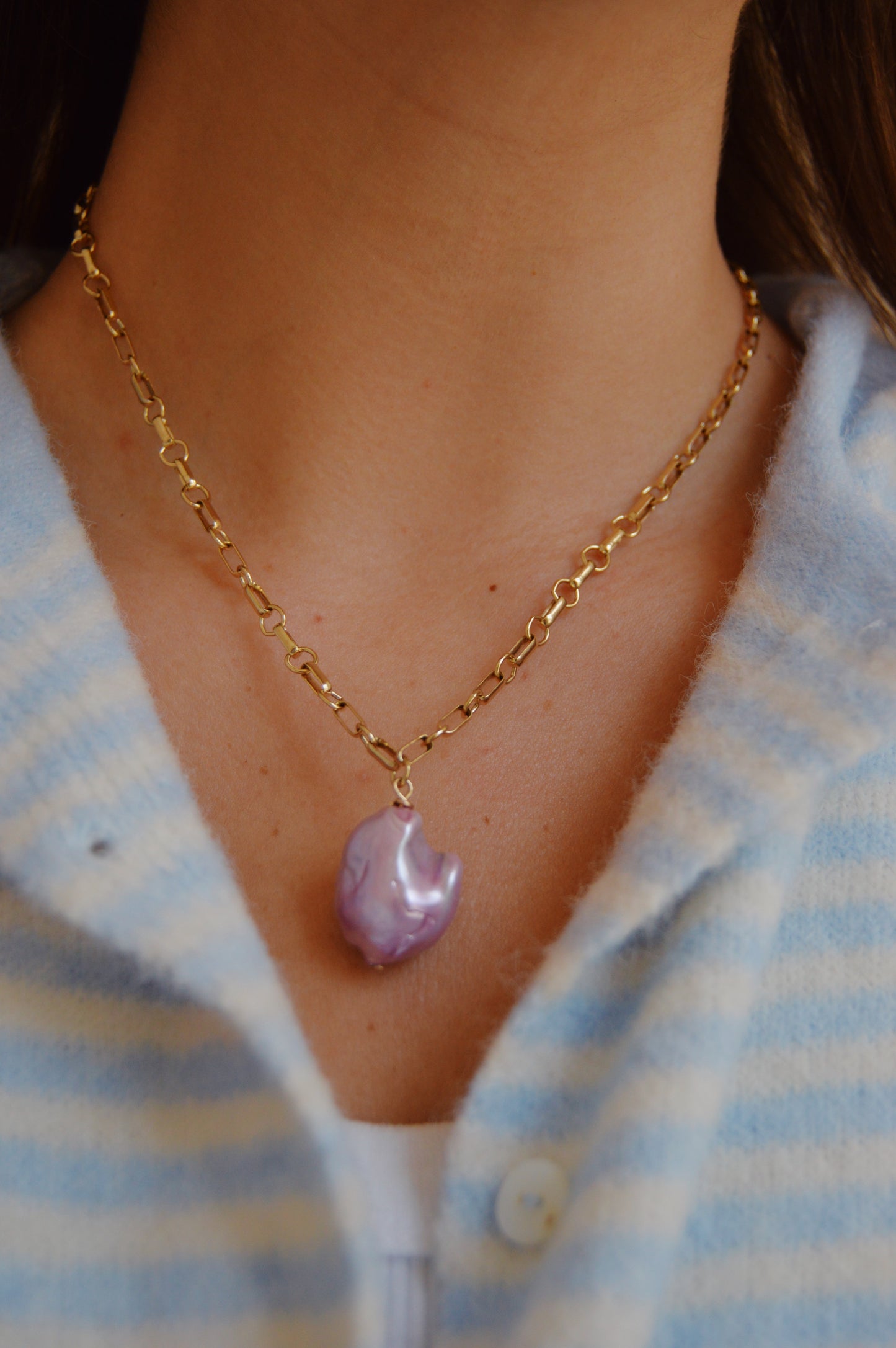A close-up of a person wearing the Lavender Pearl Drop Necklace—a 14k gold-filled chain with a purple, irregular-shaped pendant—styled over a light blue and white striped button-up shirt; an elegant handcrafted accessory.