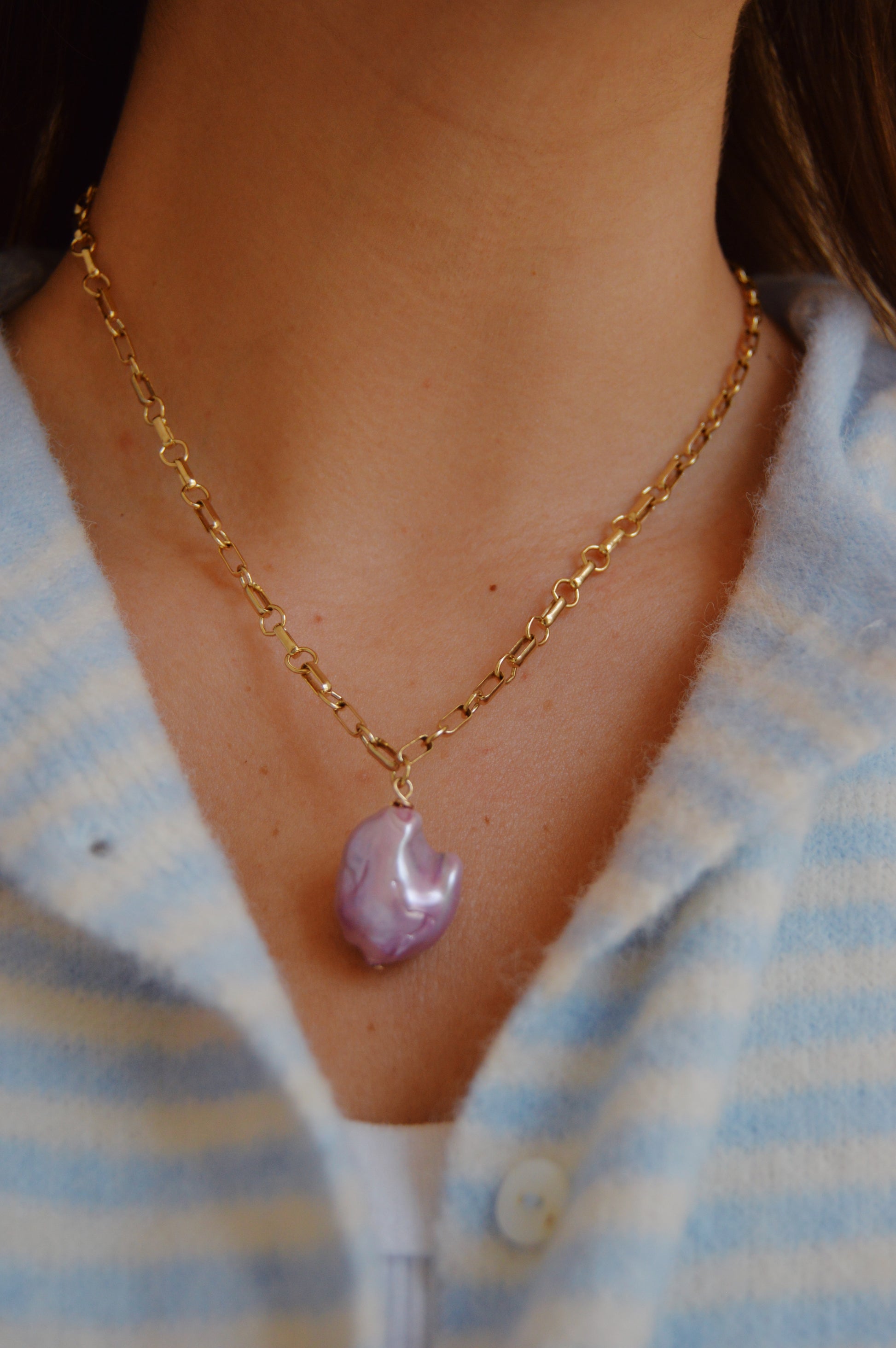 A close-up of a person wearing the Lavender Pearl Drop Necklace—a 14k gold-filled chain with a purple, irregular-shaped pendant—styled over a light blue and white striped button-up shirt; an elegant handcrafted accessory.