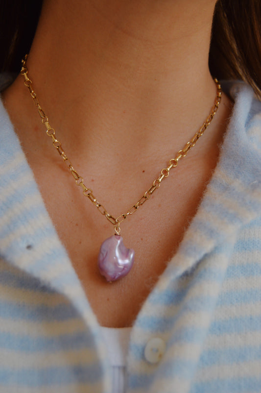 A close-up of a person wearing the Lavender Pearl Drop Necklace—a 14k gold-filled chain with a purple, irregular-shaped pendant—styled over a light blue and white striped button-up shirt; an elegant handcrafted accessory.