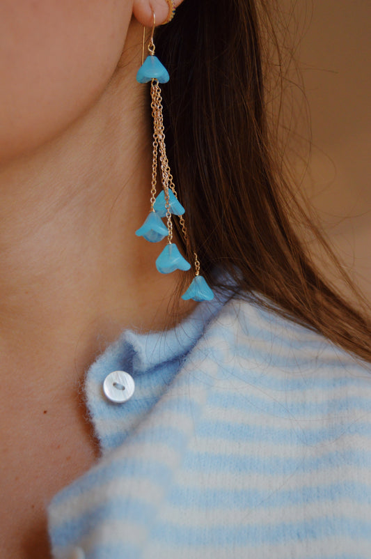 Close-up of a person wearing the Bouquet Drops- Blueberry earrings, featuring dangling blue Czech Glass Tulips. The handcrafted gold jewelry stands out against long brown hair and a blue-and-white striped fuzzy top with a white button.