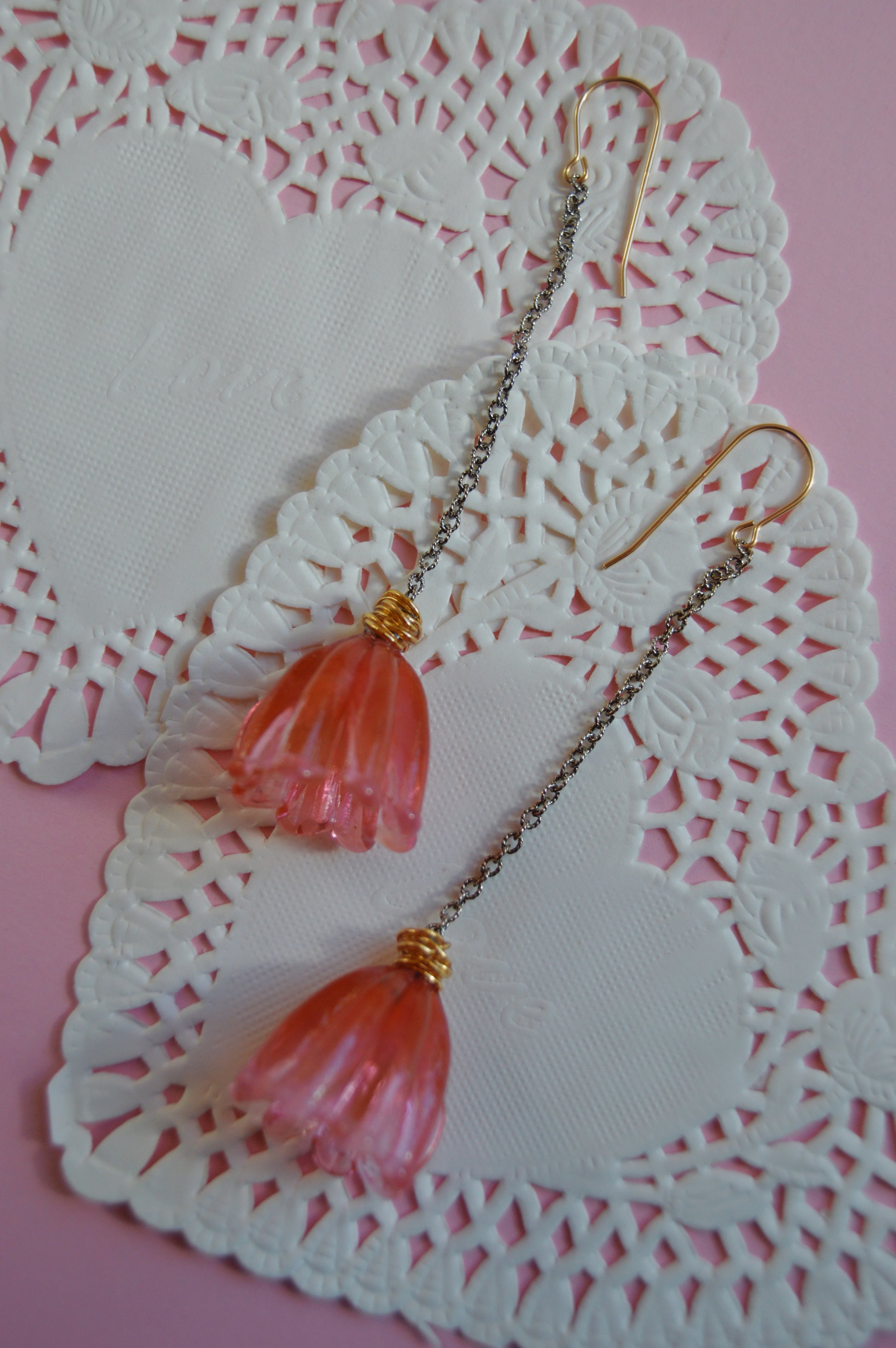 The Valentine Mixed Metal Tulips are two pink flower-shaped drop earrings with gold hooks and delicate chains, displayed on white heart-shaped doilies against a pink background.
