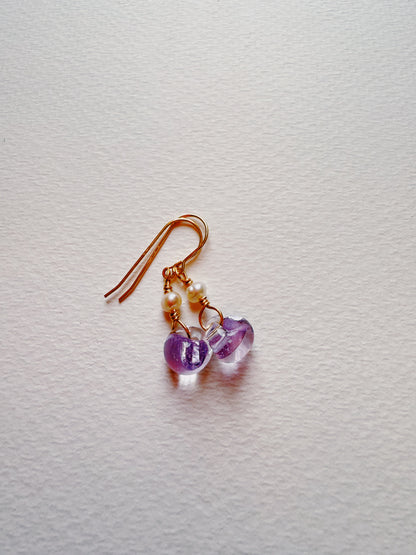 Bubble Minis gold wire earrings with two small white freshwater pearls and two translucent purple glass heart-shaped beads, shown on a textured white surface.
