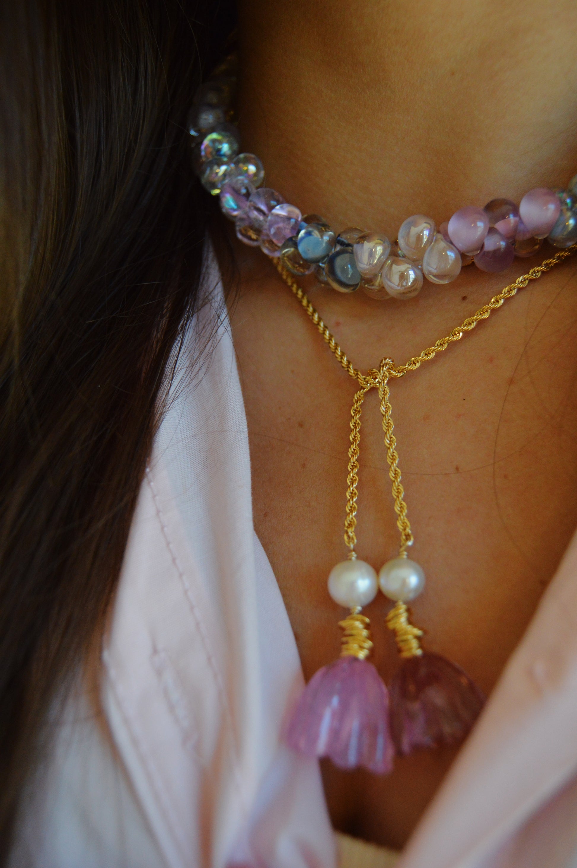 Close-up of a person's neck wearing the Valentine Bubble Glass Necklace- Sweetheart, featuring shiny iridescent purple and blue beads paired with a gold necklace adorned with pearl and pink flower-shaped pendants over a light pink top.