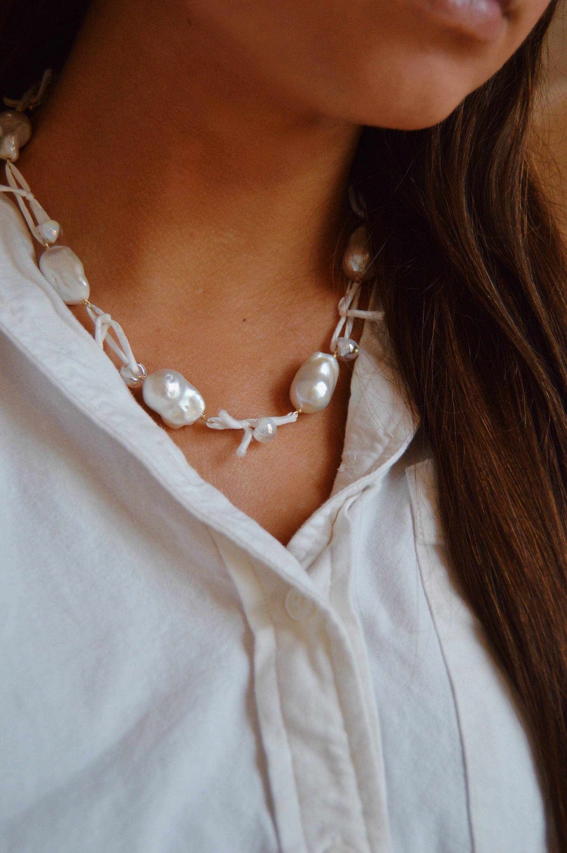 A close-up shows a person wearing the Silk and Pearl Layer - White: a chunky, handmade necklace with irregular freshwater pearls on white silk. The person pairs it with a white button-up shirt, and their long brown hair is visible on one side.