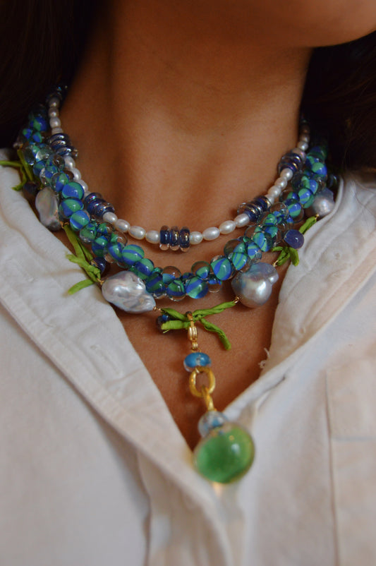 A close-up of someone wearing Stack #4—blue, green, and pearl layered necklaces with mixed beads and a gold pendant—styled with a white shirt. Ideal for your giveaway stacks or to snag now at 20% off.