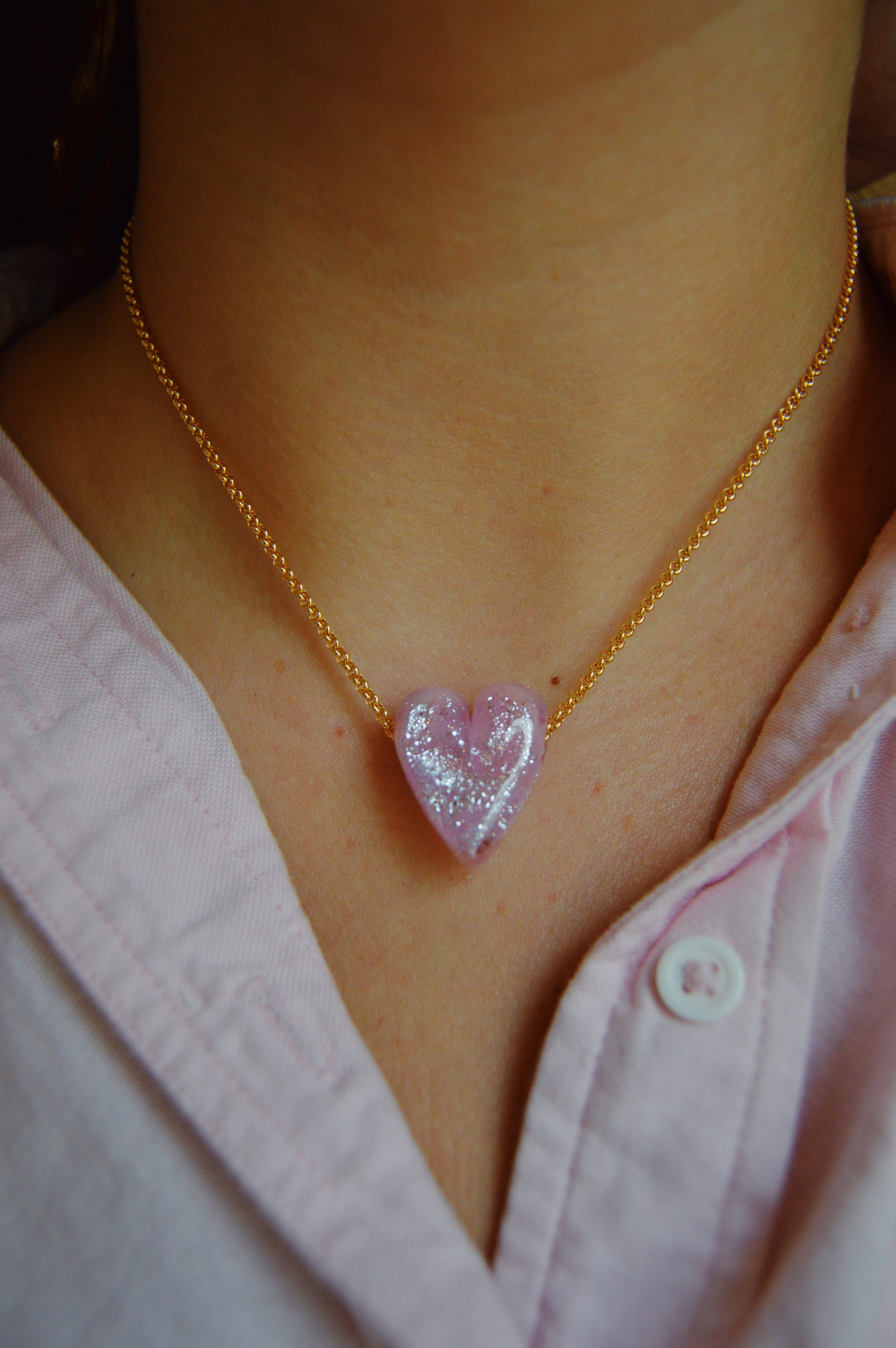 Someone wearing a pink shirt has a Glass Heart Necklaces- Pinks—a gold-filled chain with a sparkling, handmade lampwork glass heart pendant—around their neck.