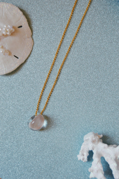 The Grand Bubble Single - Nude, a gold beaded necklace with cultured freshwater pearls and 14k gold filled accents, featuring a clear stone pendant, is displayed on a sparkly blue surface beside a white sand dollar and coral.