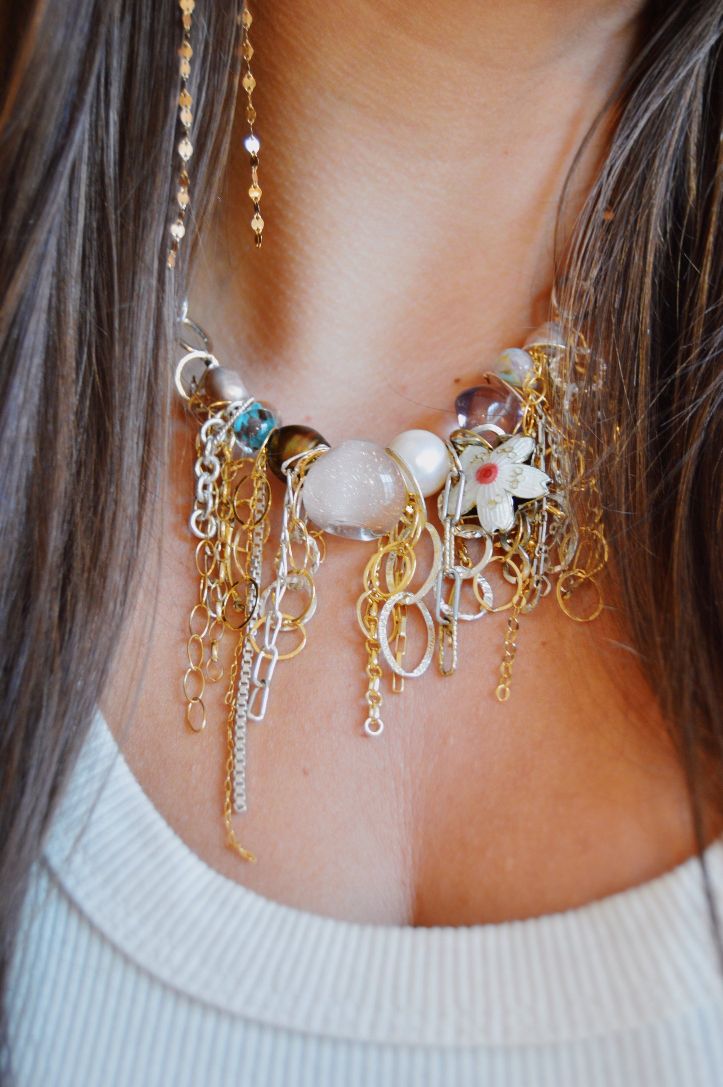 A close-up of a woman wearing the Mixed Metal Chain Layer, a chunky necklace featuring gold and silver chains, beads, pearls, a white flower charm, and decorative details. She pairs it with a white ribbed top.