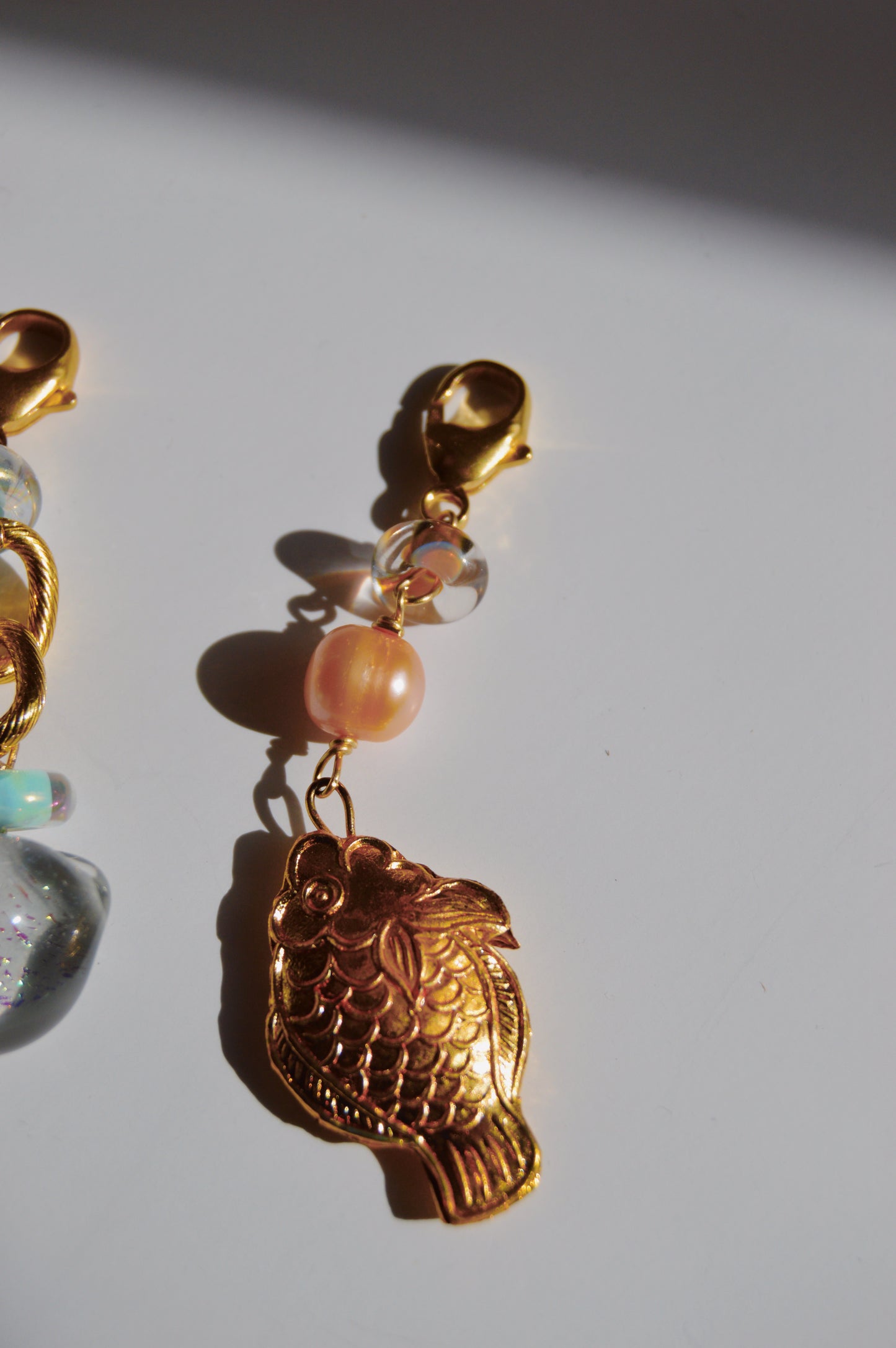 The Fish and Pearl Charm features a Cloisonne fish hanging from a lobster clasp, accented with a pink cultured freshwater pearl and clear round beads, casting a delicate shadow on a white surface.