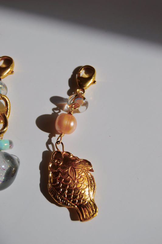 The Fish and Pearl Charm features a Cloisonne fish hanging from a lobster clasp, accented with a pink cultured freshwater pearl and clear round beads, casting a delicate shadow on a white surface.