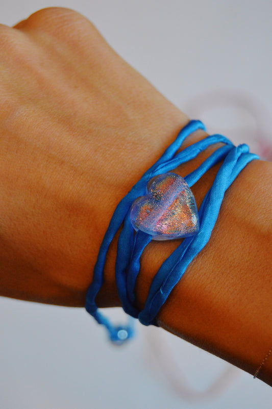 A hand models the Silk Glass Heart- Cobalt: an artisan bracelet featuring a sparkling, handmade lampwork glass heart-shaped charm on a blue silk ribbon, stylishly wrapped multiple times around the wrist.