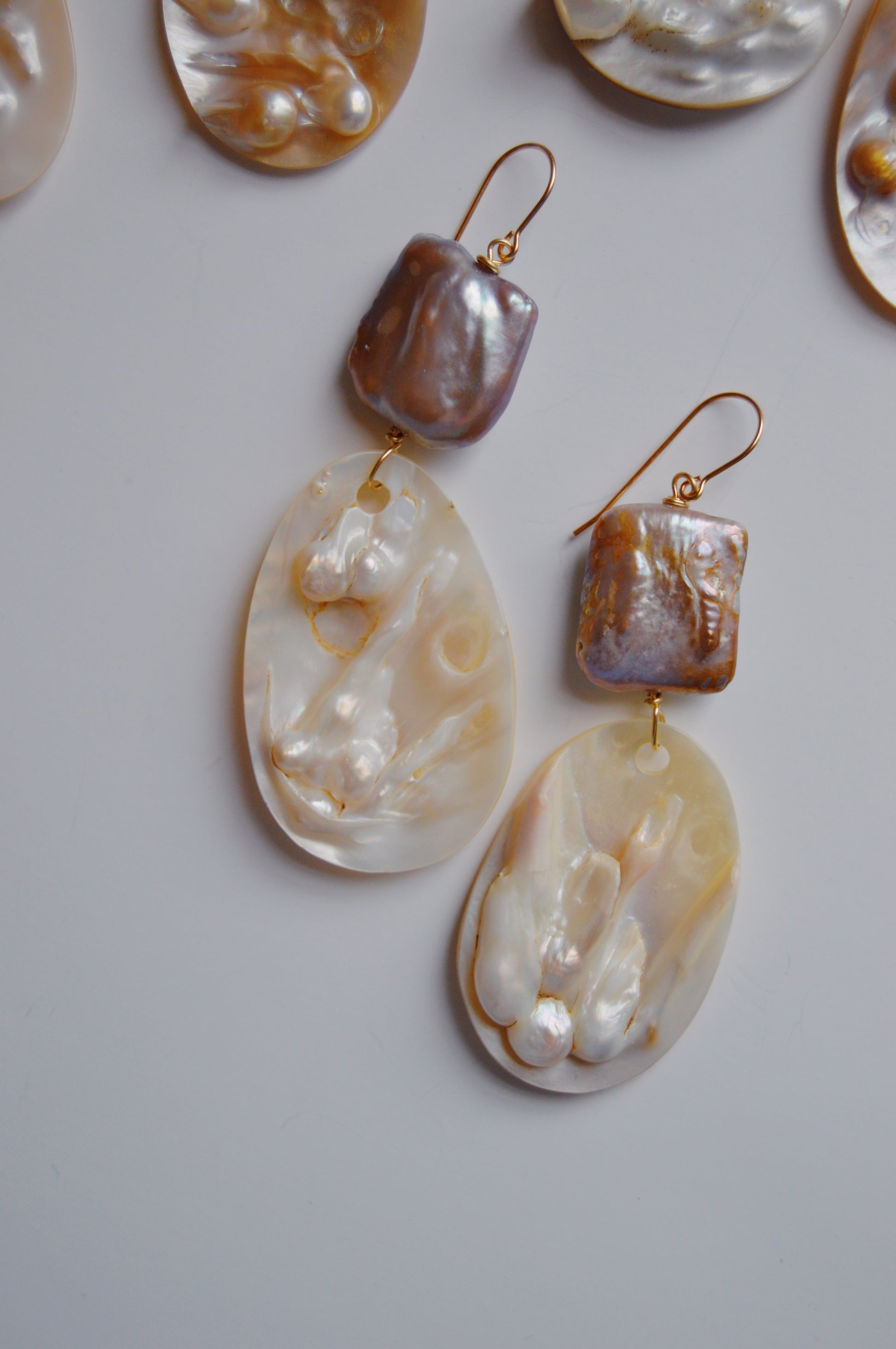 Angel Shell Drops: Handcrafted dangle earrings with 14k gold-filled hooks, featuring a square iridescent bead and an oval, wavy-textured pearlescent pendant—displayed on a white background.