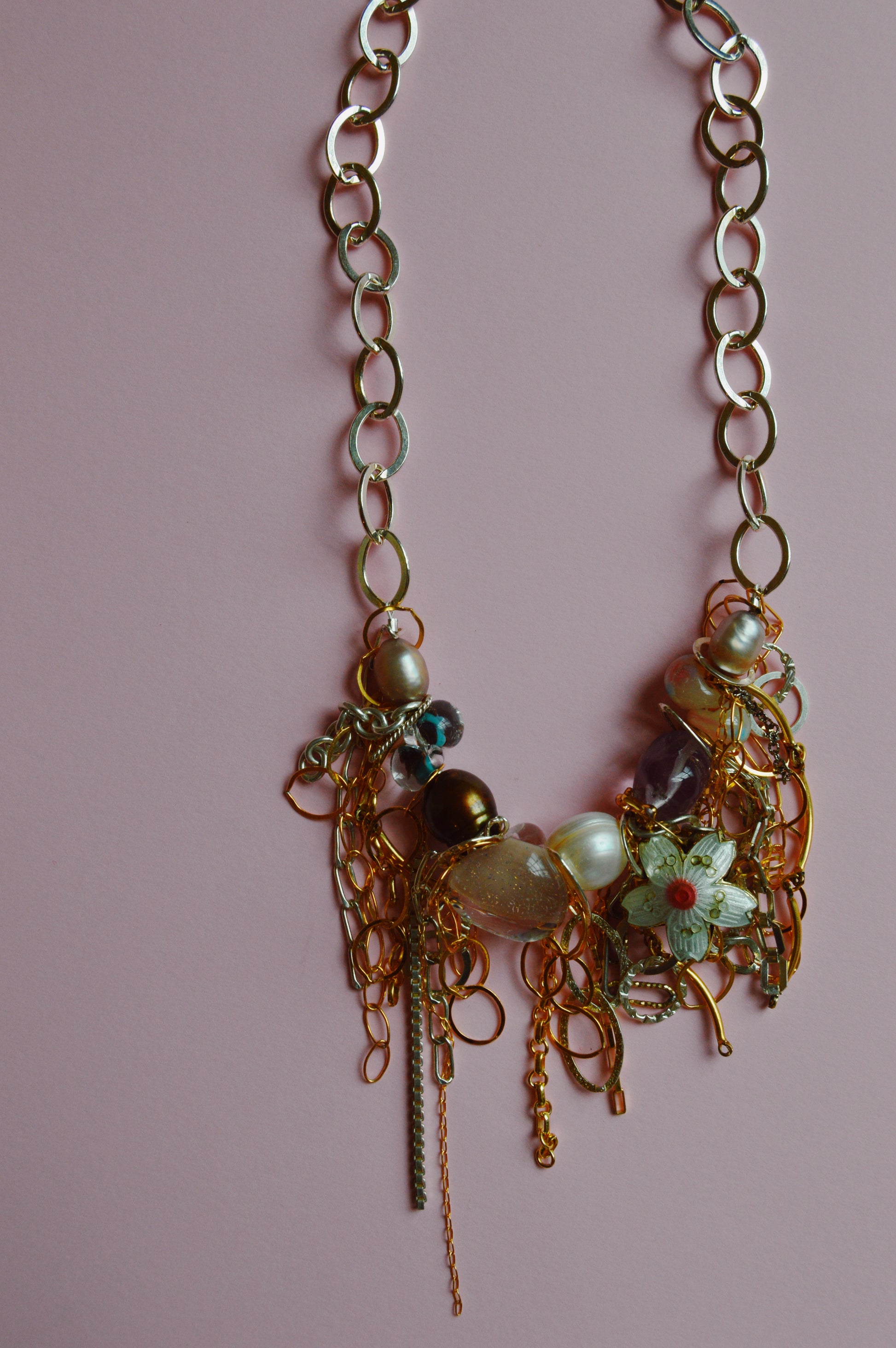 The Mixed Metal Chain Layer showcases large gold links adorned with beads, pearls, and floral charms, set against a pink background.