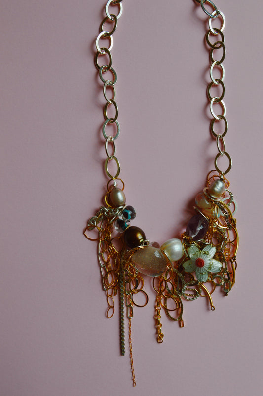 The Mixed Metal Chain Layer showcases large gold links adorned with beads, pearls, and floral charms, set against a pink background.