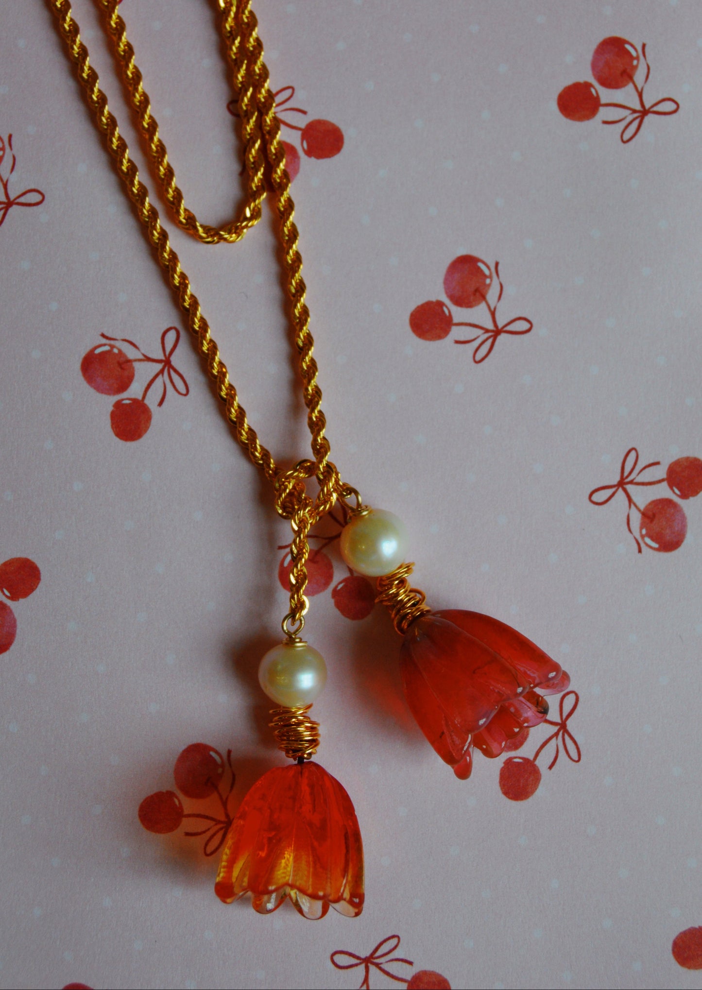 The Tulip Wrap Necklace-Cherry on Top is a gold twisted chain with two pendants—one has orange flower-shaped beads and the other round pearl-like beads, shown on a pink cherry-patterned background.
