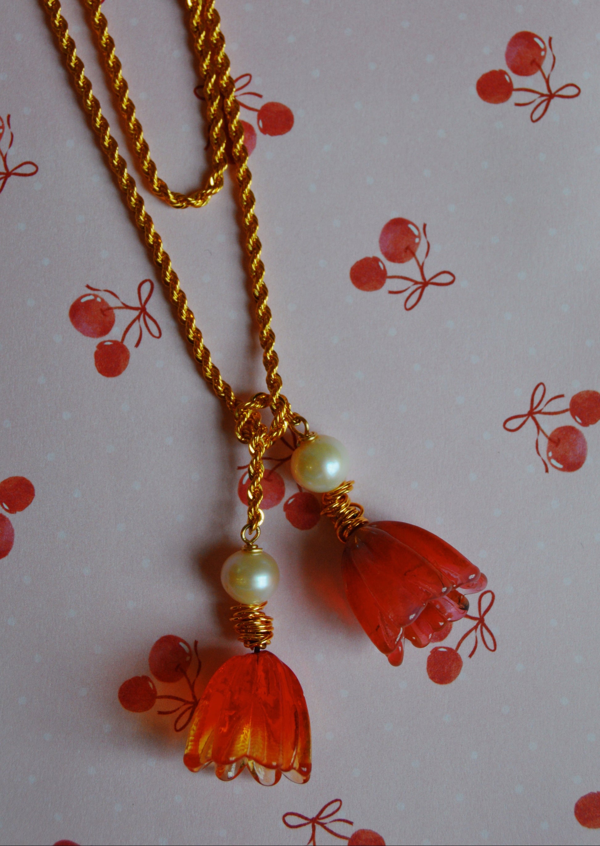The Tulip Wrap Necklace-Cherry on Top is a gold twisted chain with two pendants—one has orange flower-shaped beads and the other round pearl-like beads, shown on a pink cherry-patterned background.