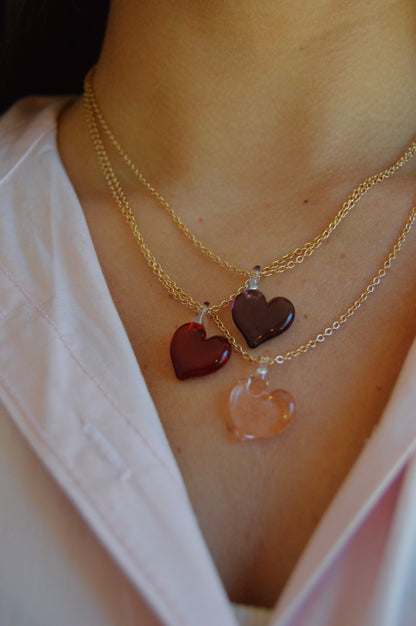 A close-up of a person in a light pink shirt wearing the Flat Glass Heart Layer: two gold filled chain necklaces, each with handblown glass heart pendants in red, dark red, and light pink.