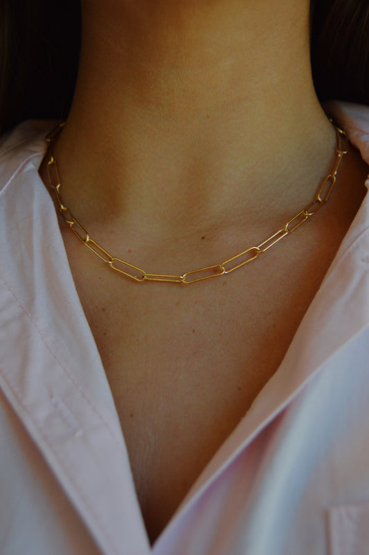 A close-up of a person wearing a light pink collared shirt and the Paperclip Layer, a delicate gold chain necklace with elongated links, around their neck.