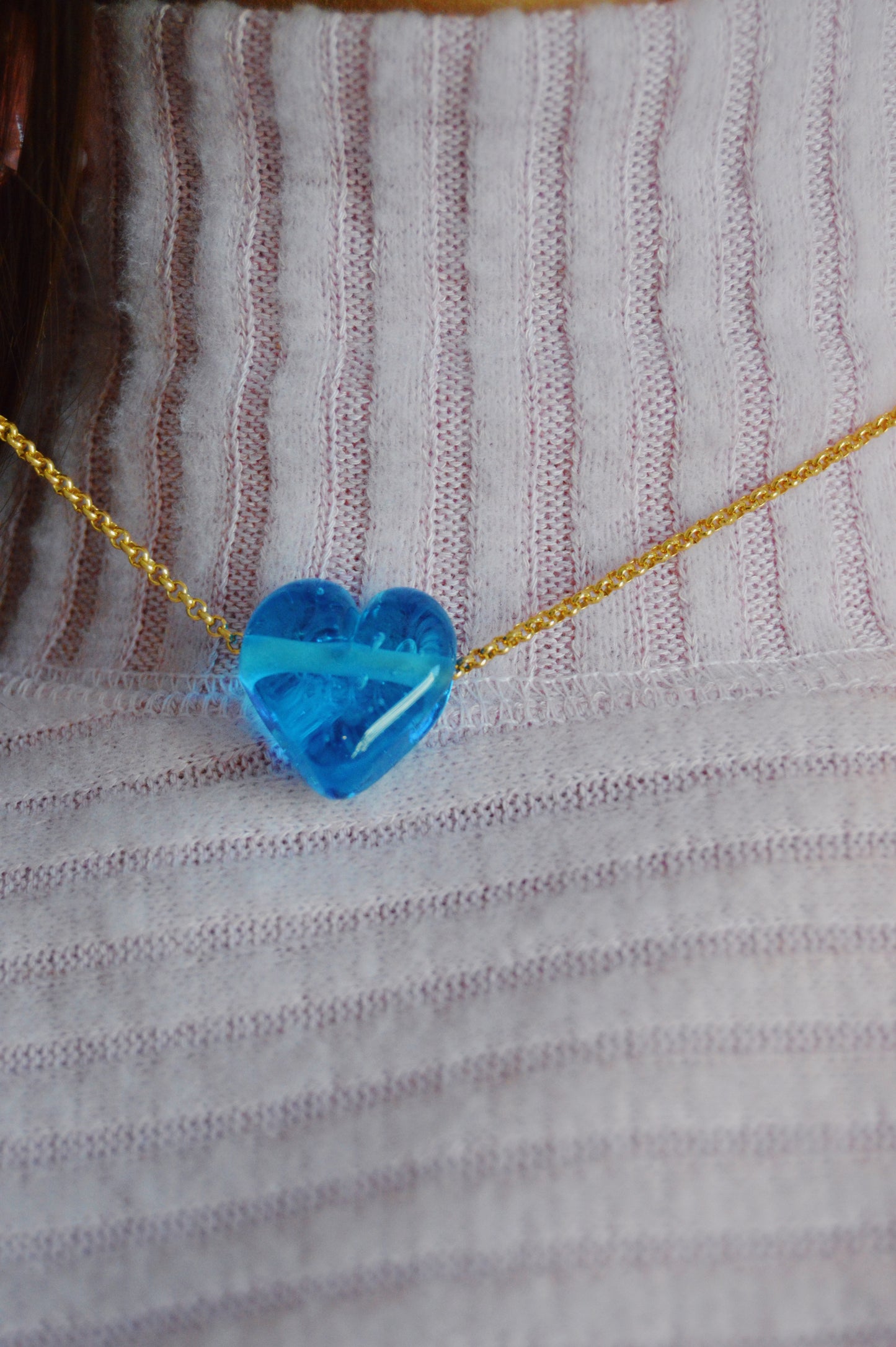 The Glass Heart Necklaces- Blues features a handmade lampwork glass heart pendant on a gold-filled chain, styled over a light pink, ribbed shirt.