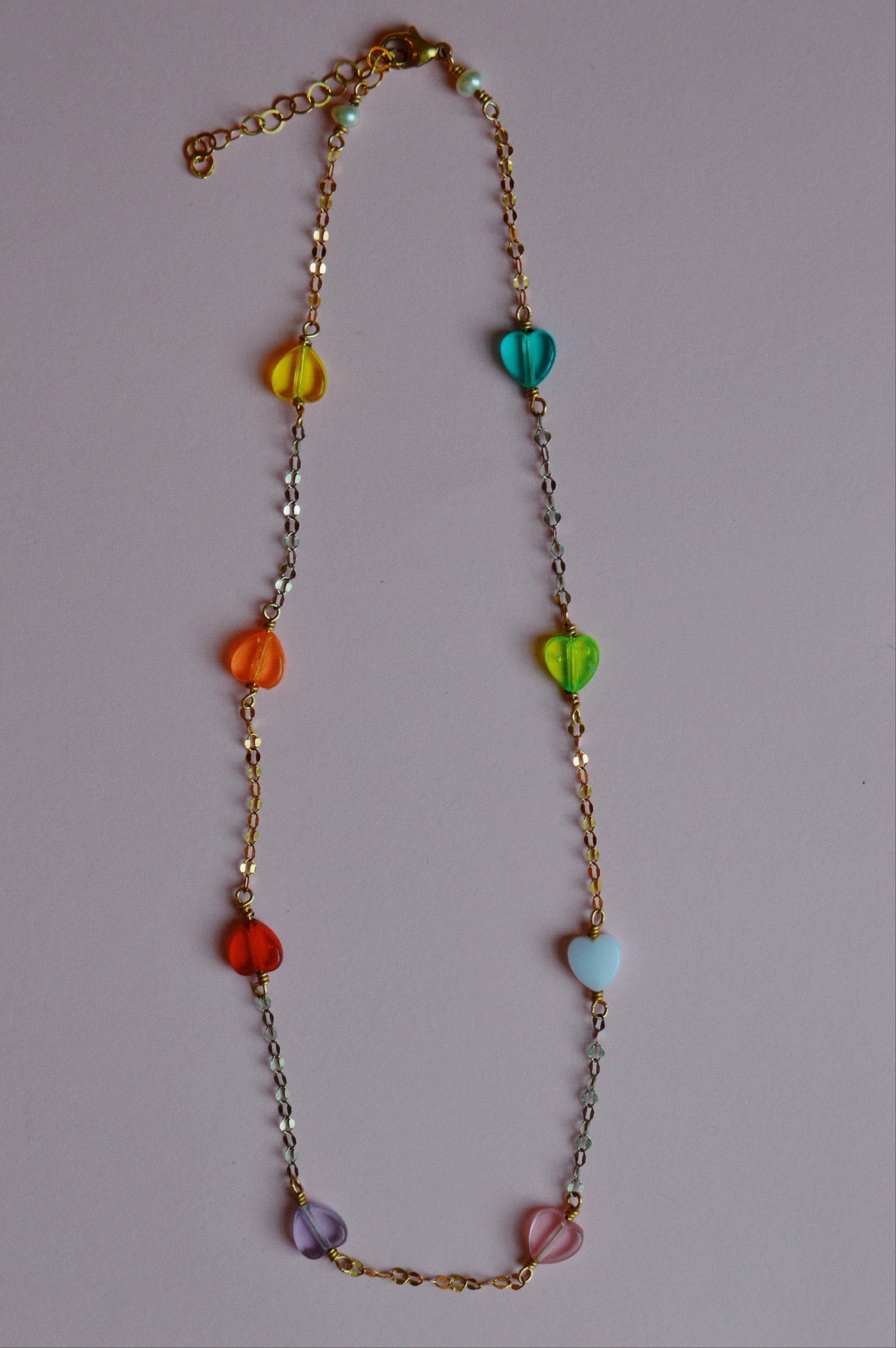 The Valentine Acrylic Heart Layer necklace is a 14k gold-filled chain with six colorful acrylic heart beads, shown on a plain light pink background.