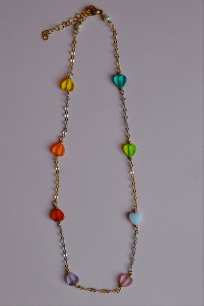 The Valentine Acrylic Heart Layer necklace is a 14k gold-filled chain with six colorful acrylic heart beads, shown on a plain light pink background.