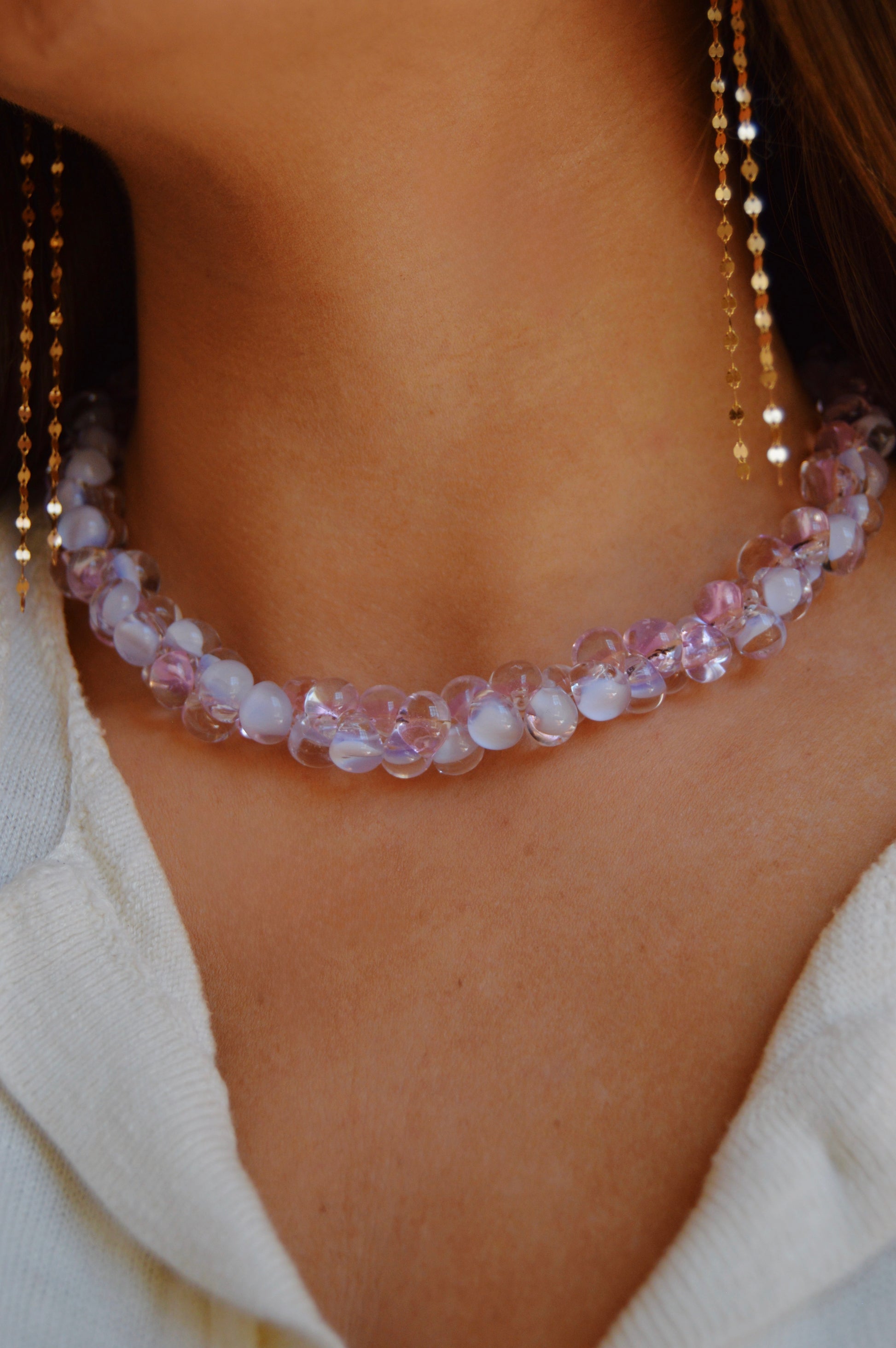 A close-up of a person wearing the Bubble Glass Necklace- Pink and Lavender with translucent pink and lavender beads, paired with gold-filled earrings and a white textured top.