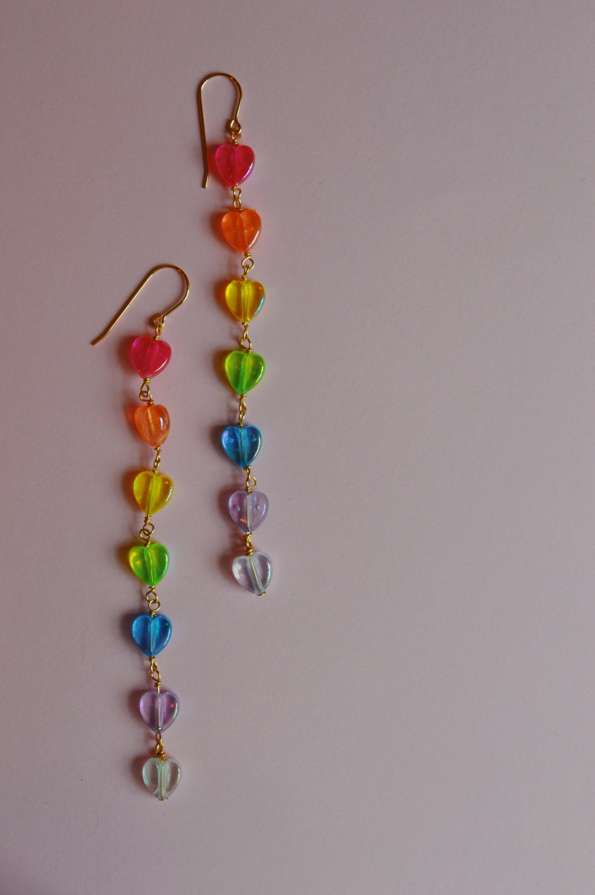 The Valentine Rainbow Acrylic Drops are two dangling earrings with small, translucent, rainbow-colored acrylic heart beads and 14k gold filled findings, shown on a light pink background.