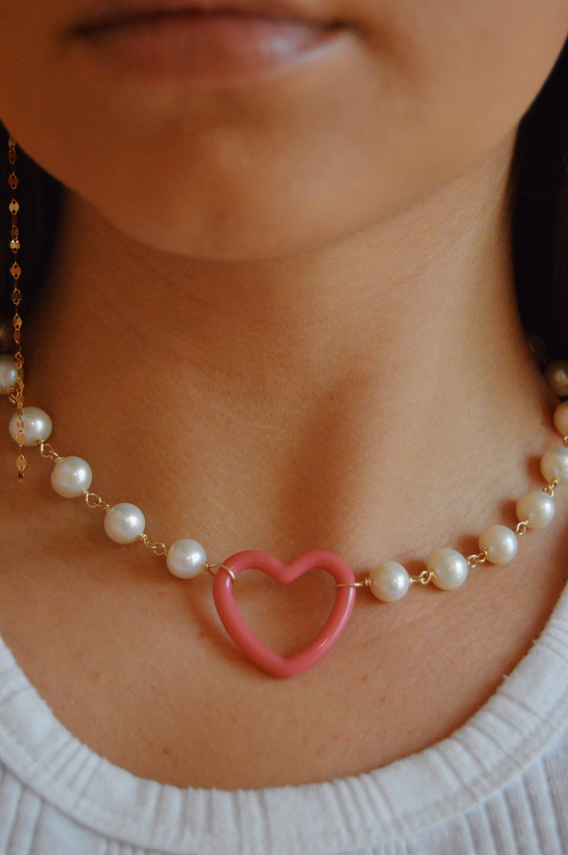 A close-up of a person wearing the "Open Heart on Pearl Chain" necklace, featuring a pink acrylic heart charm and freshwater pearls, paired with a white top.