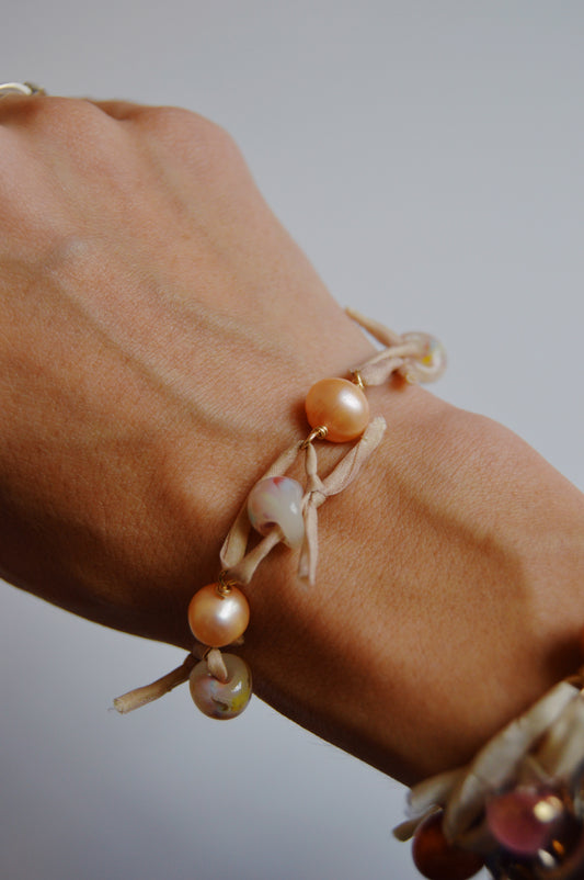 A close-up shows a wrist wearing the Silk and Pearl Bracelet- Peachy Puka, crafted from light ribbon with round peach and white beads reminiscent of freshwater pearls. The background is plain and out of focus.