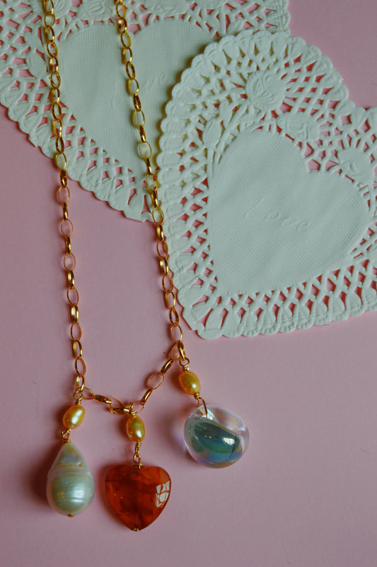 The Charm Necklace features a gold chain with three heart-shaped pendants in red, blue, and pale green, showcased on a pink background next to white, heart-shaped paper doilies.