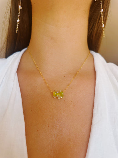 A close-up of a woman wearing the Dainty Donut Layer: a delicate 14K gold chain necklace with three greenish-yellow gemstones, paired with a white V-neck top and pearl-accented gold earrings, finished with a secure gold-filled clasp.
