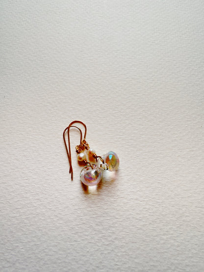 The Bubble Minis are delicate dangle earrings featuring iridescent glass beads, freshwater pearls, and gold filled findings, displayed on a textured white surface.