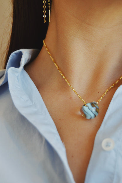 Close-up of a person in a light blue collared shirt wearing the Grand Bubble Single - Funky Fish necklace, featuring a 14k gold filled chain and round blue and white marbled pendant. The focus is on the necklace and pendant.