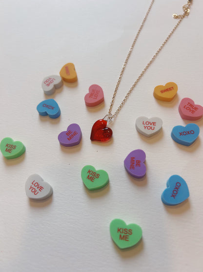 The Flat Glass Heart Layer, a 14k gold necklace with a handblown glass heart pendant, is displayed on a white surface surrounded by colorful candy hearts featuring messages like “KISS ME,” “LOVE YOU,” “XOXO,” and “BE MINE.”.