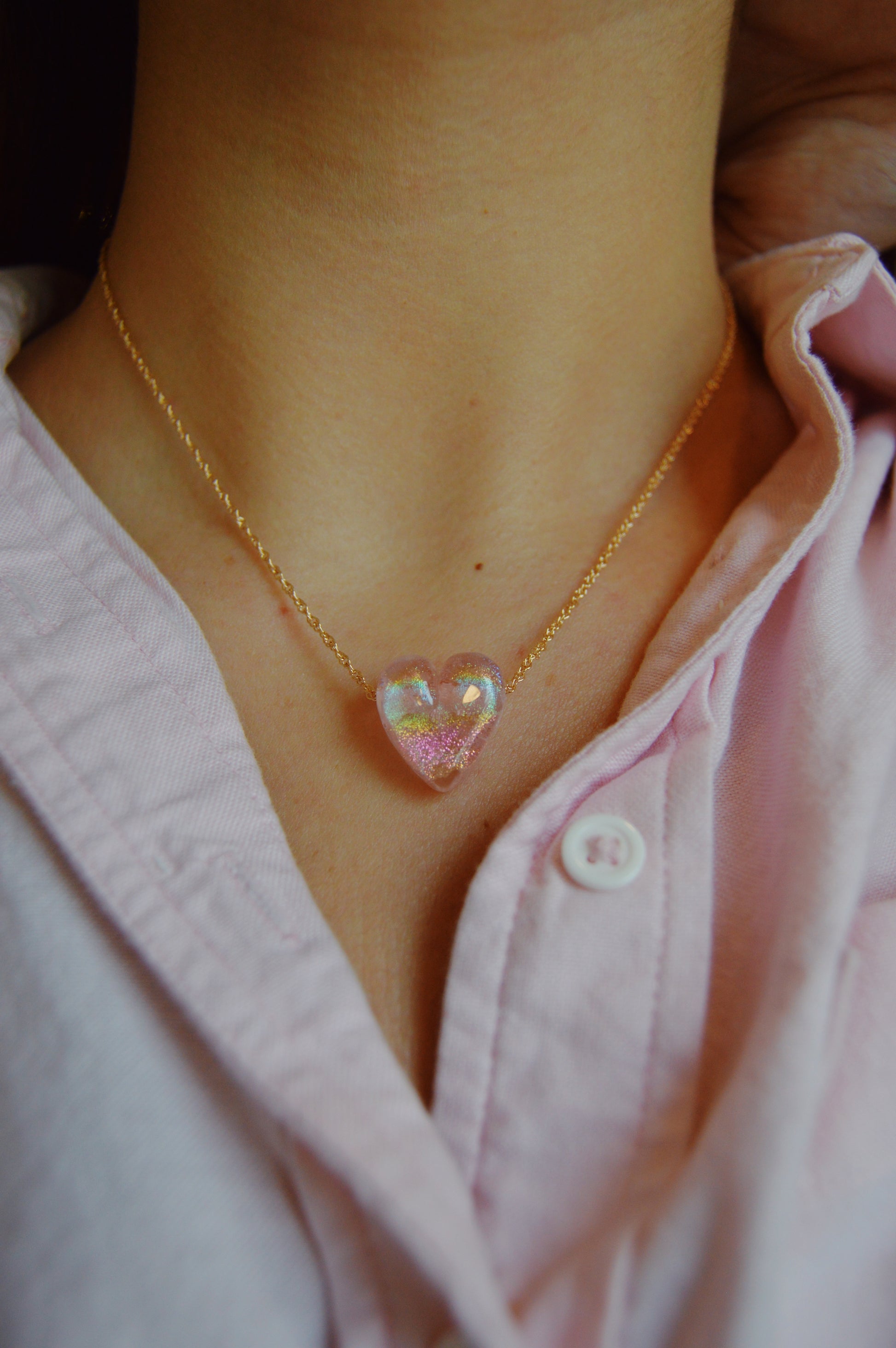 A person wears a pale pink shirt with a Glass Heart Necklaces- Pinks pendant featuring iridescent handmade lampwork glass on a gold-filled chain around their neck.