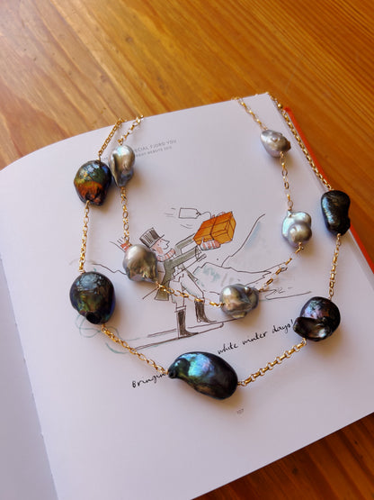 The Silver Organic Pearl Layer, a chain necklace with large multicolored baroque pearls, is displayed on an open book with an illustration of a person holding an orange gem, atop a wooden surface.