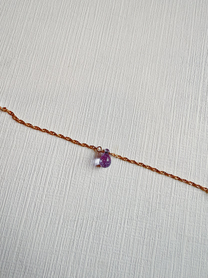 The Dewdrop Bracelet, a thin, twisted 14k gold filled adjustable chain with a small purple and clear glass bead charm, rests on a textured white surface.