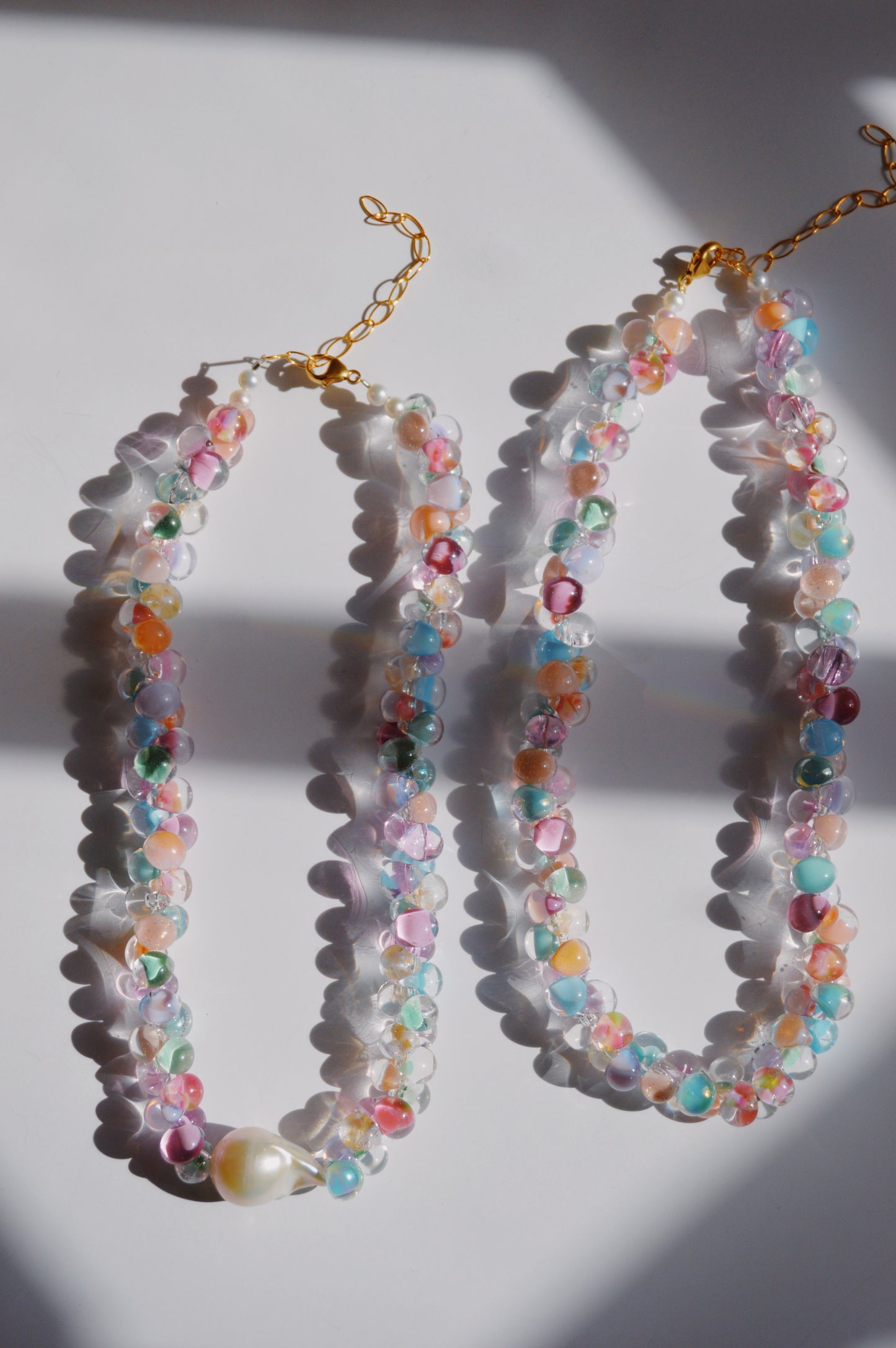The Bubble Glass Necklace—Spring Mix features colorful handmade glass beads with gold chain clasps, arranged on a white surface in sunlight and shadow beside a freshwater pearl necklace with a large pearl accent.