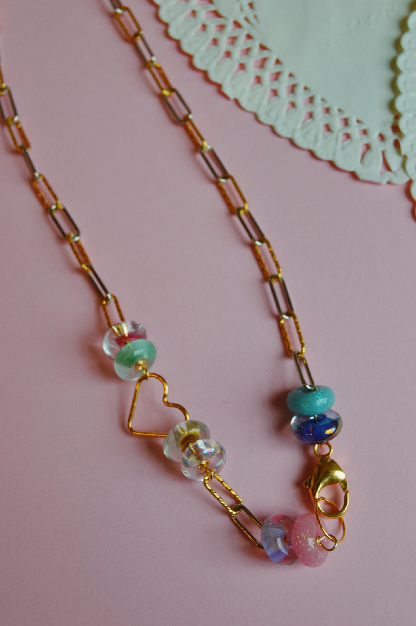 The Valentine Clasp in Front Heart Necklace, featuring colorful glass beads, is displayed on a pastel pink surface with a white paper doily partly visible in the top right corner.