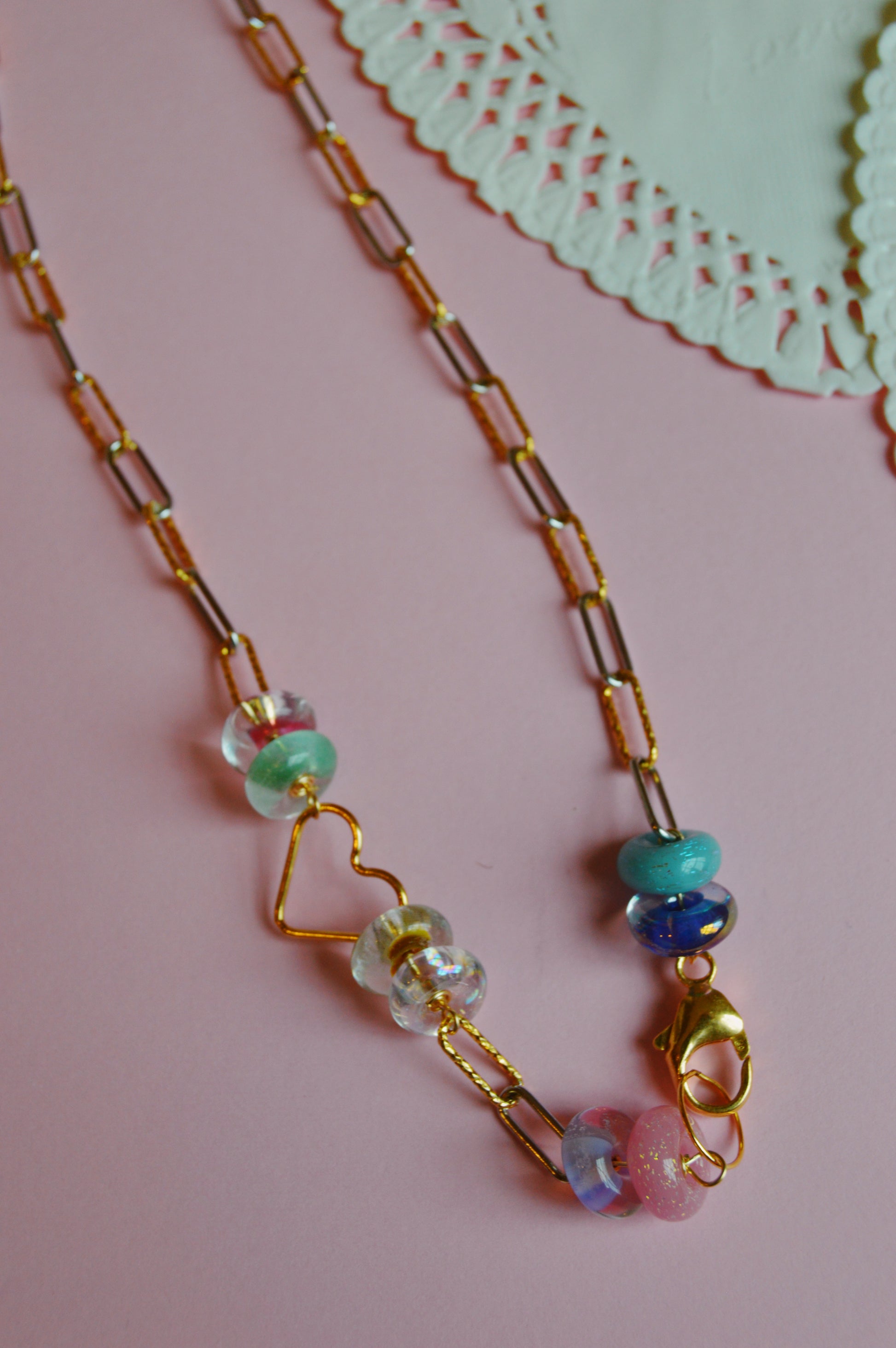The Valentine Clasp in Front Heart Necklace, featuring colorful glass beads, is displayed on a pastel pink surface with a white paper doily partly visible in the top right corner.