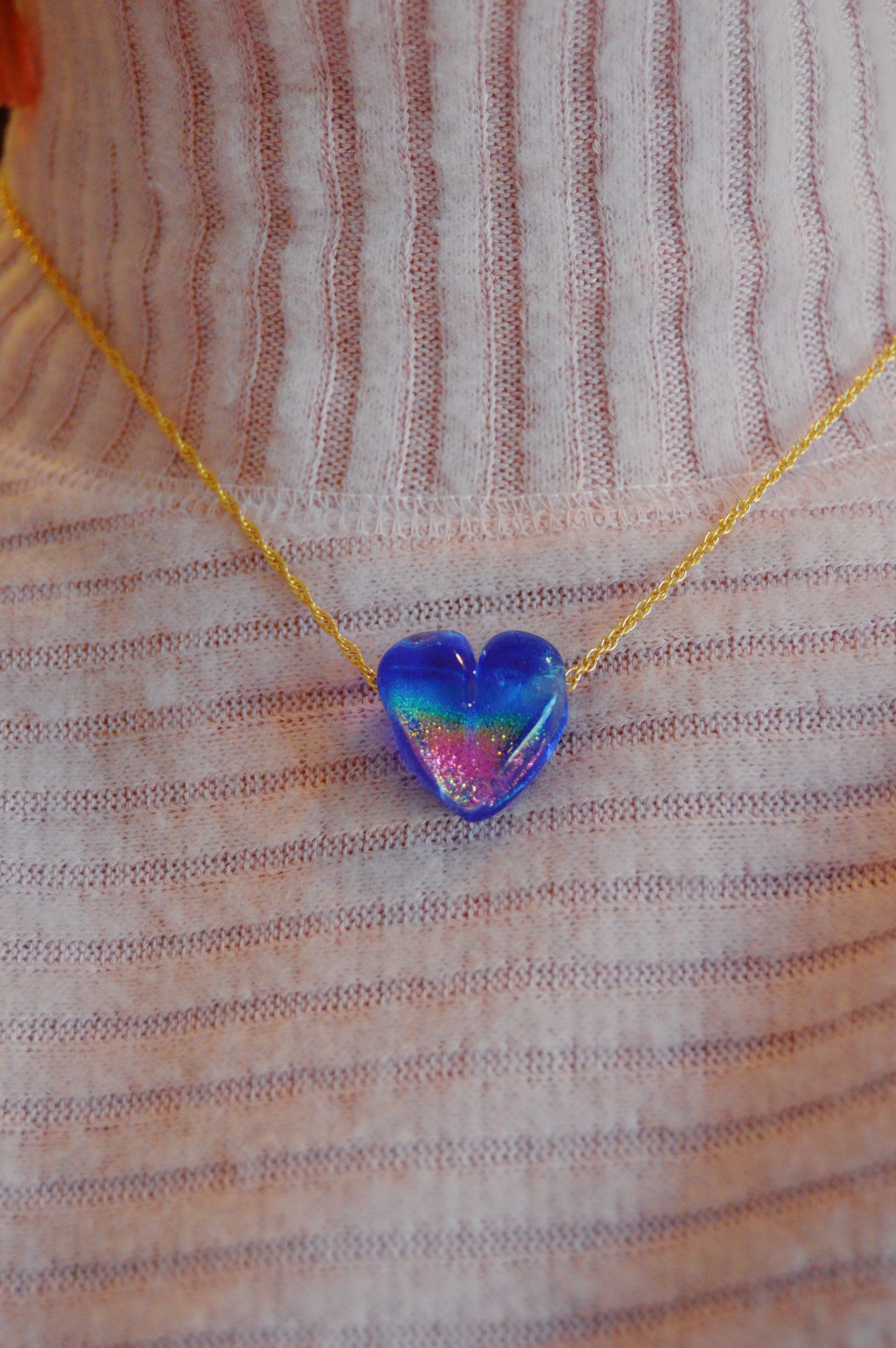 A person wears a ribbed light pink top and the Glass Heart Necklaces- Blues: a gold-filled chain with an artisan, iridescent blue handmade lampwork glass heart pendant.